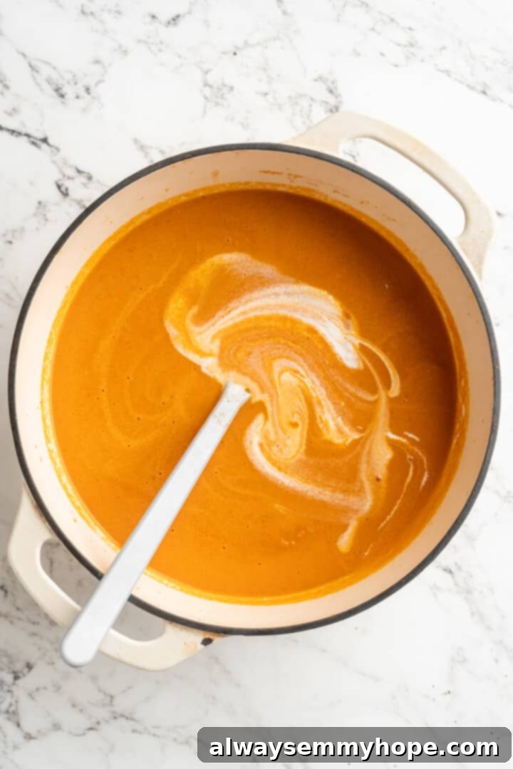 Overhead view of cashew cream being poured into the pot of smooth tomato soup, ready to be stirred in for ultimate creaminess.