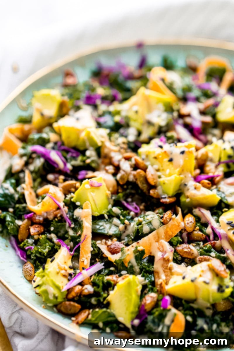 This is the BEST kale salad recipe! With tender massaged kale, avocado, carrots, and a crunchy seed blend tossed in creamy tahini dressing. Close up of kale salad topped with avocado, carrots, seeds, and dressing in a large serving bowl.