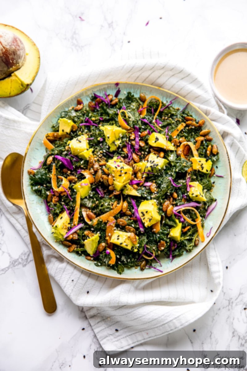 This is the BEST kale salad recipe! With tender massaged kale, avocado, carrots, and a crunchy seed blend tossed in creamy tahini dressing. Overhead view of kale salad topped with avocado, carrots, seeds, and dressing in a large serving bowl, next to a spoon and a small bowl of tahini dressing.