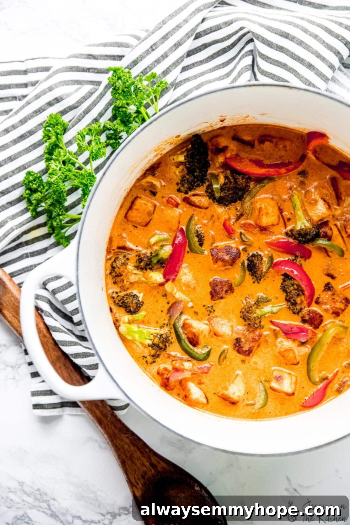Overhead view of Thai red curry in enamel Dutch oven with wooden spoon, tea towel, and sprig of parsley
