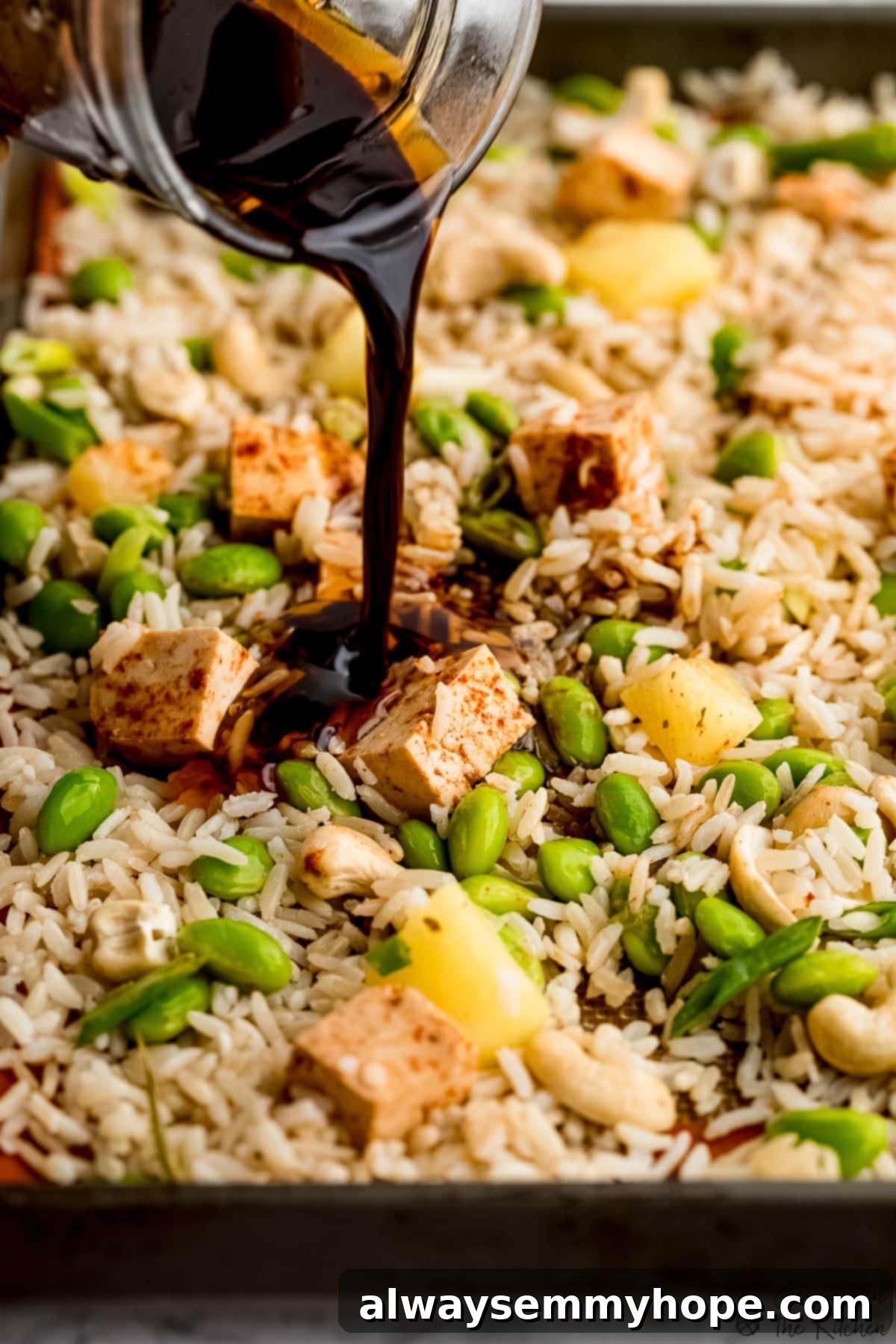 The crucial step: generously coating ingredients on the sheet pan with our irresistible fried rice sauce. A hand pouring a rich, savory sauce over a sheet pan filled with brown rice, green onions, pineapple, edamame, and tofu.