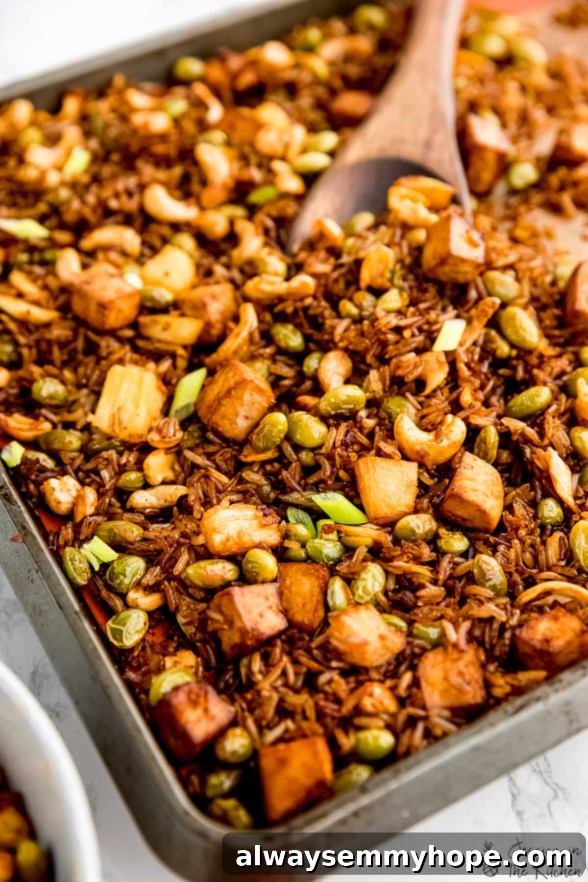 Feast your eyes on this colorful sheet pan of vegan fried rice, featuring tofu, pineapple, and cashews, about to transform in the oven! A close-up of a sheet pan filled with beautifully mixed vegan fried rice, showing crispy grains, cubed tofu, bright pineapple, and green onions.