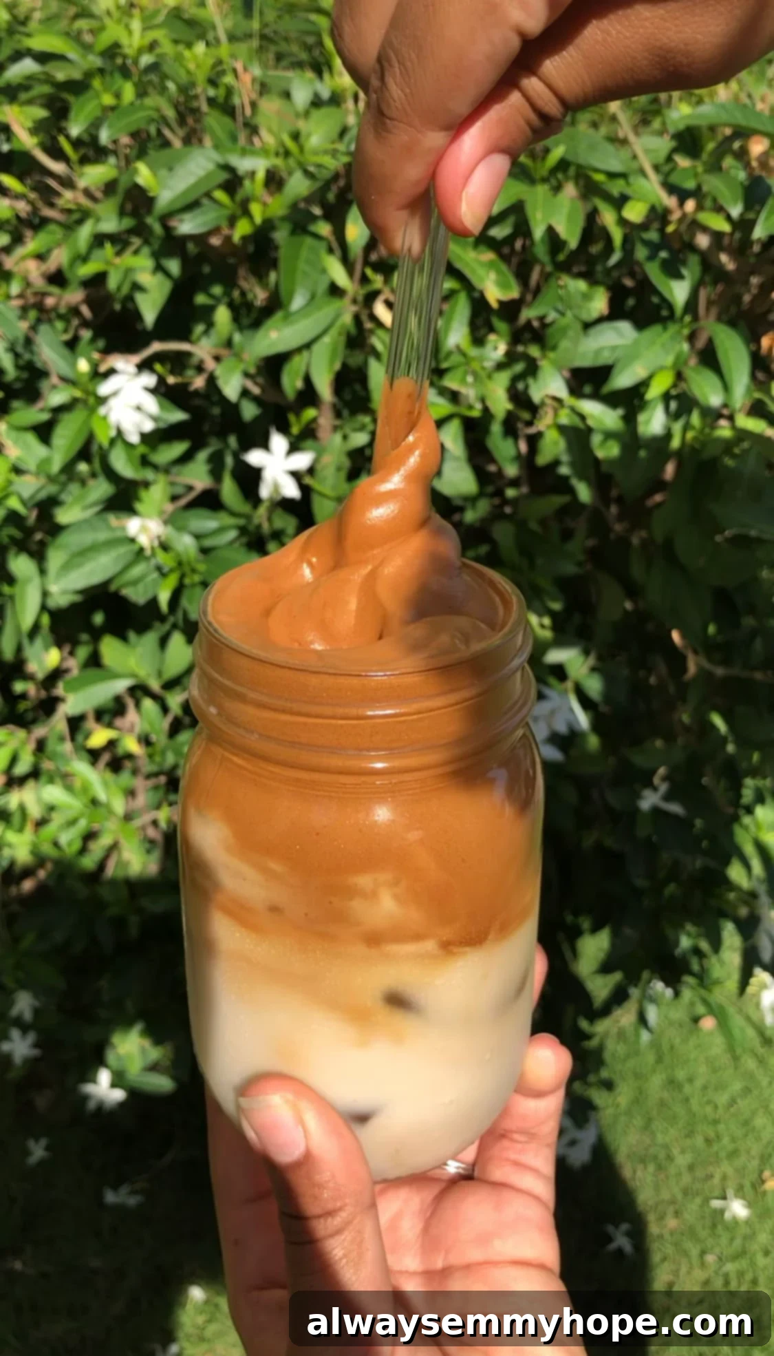 Dalgona Kahve Nasıl Yapılır Püf Noktalarıyla 2 A tall mason jar filled with a frothy whipped coffee topping over milk, with a glass straw gently swirling the layers together, highlighting its creamy texture.