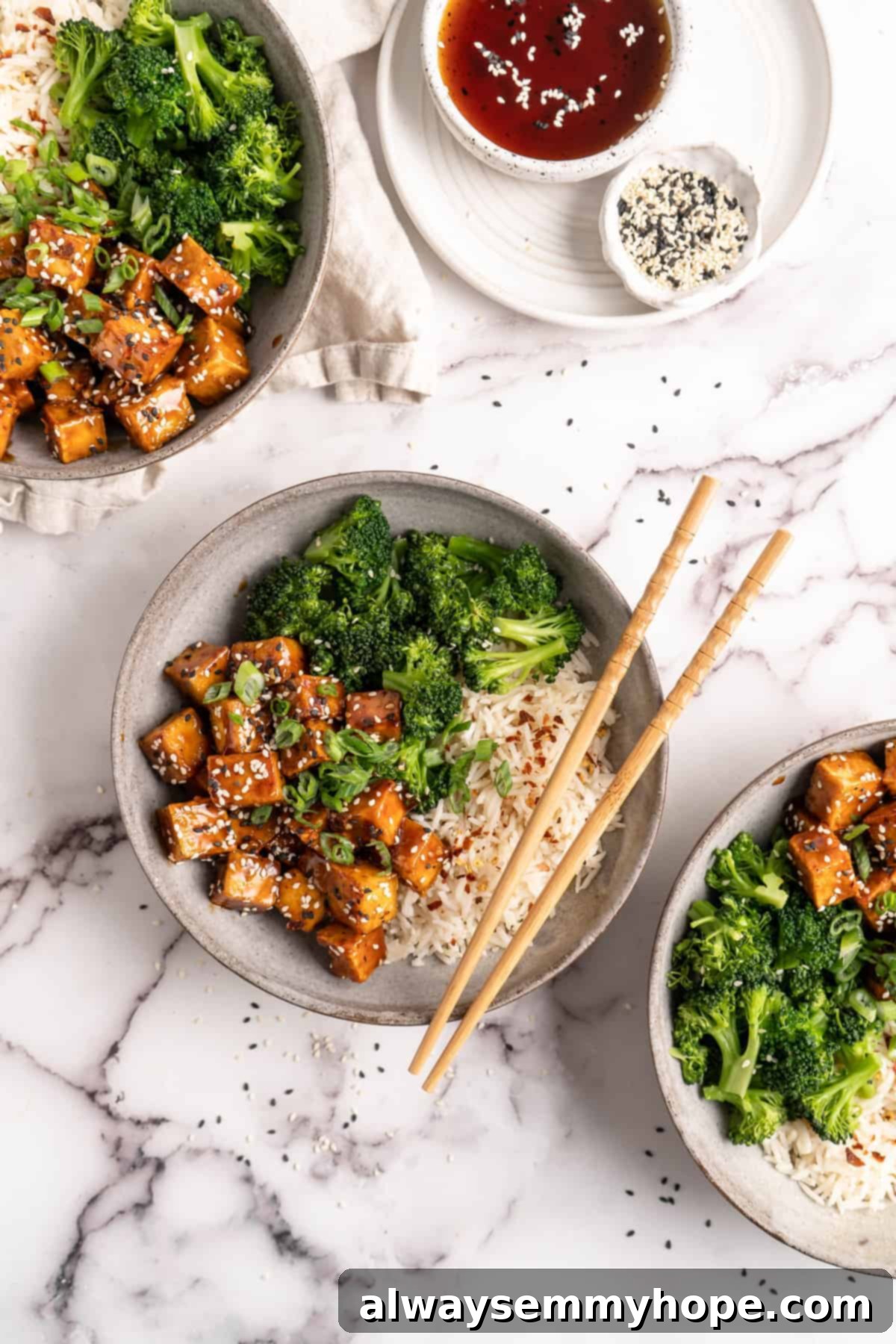 Three bowls of General Tso Tofu with white rice and broccoli, garnished with sesame seeds and green onions
