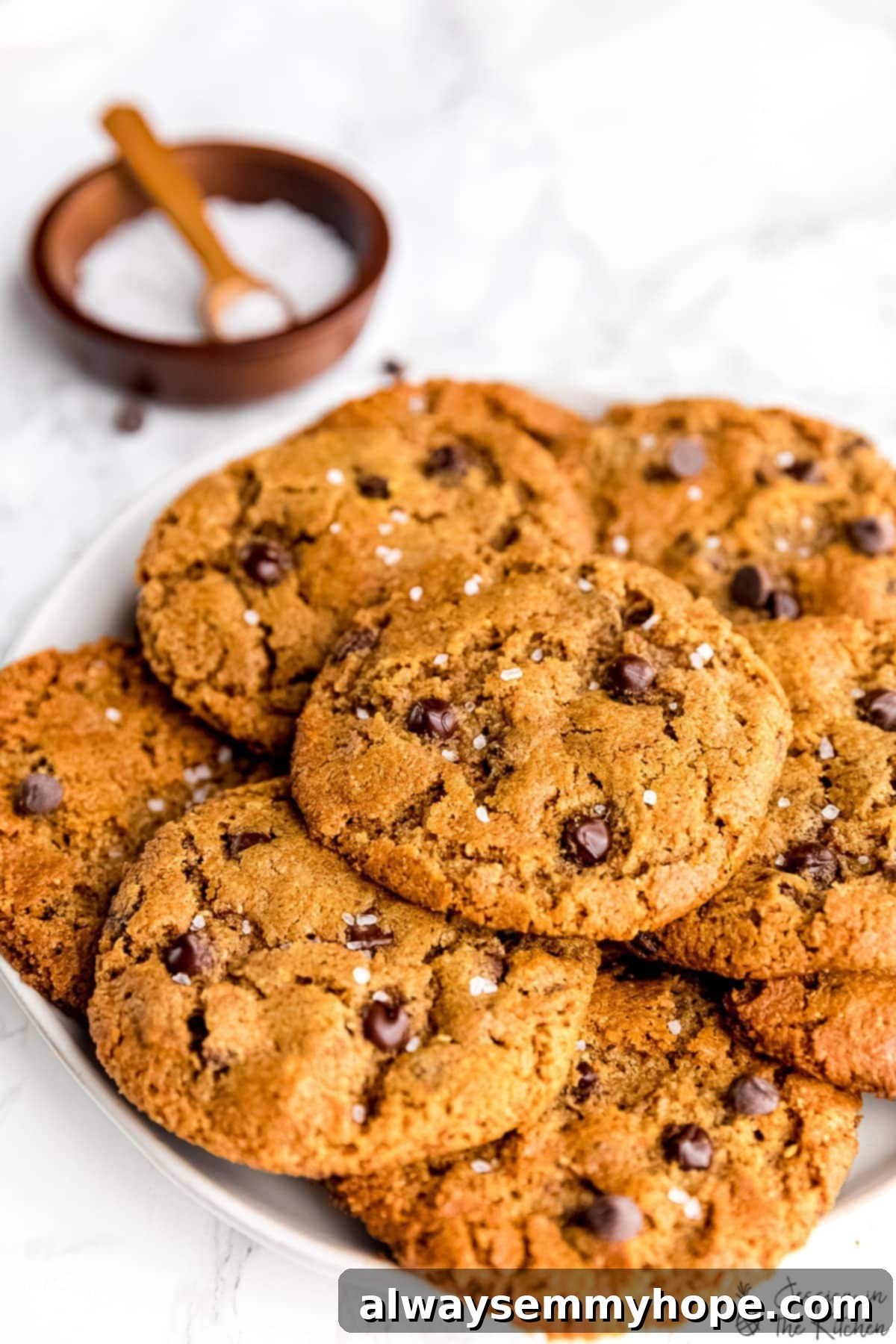 This is the best vegan chocolate chip cookies recipe! With only 5 ingredients, these vegan cookies are soft-baked, chewy, and EASY to make! Close up of vegan chocolate chip cookies piled on a white plate with a small bowl of sea salt in the background.