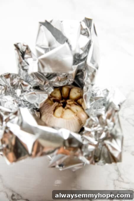 Garlic head being wrapped in foil for roasting. Hands carefully wrapping a prepared garlic head tightly in aluminum foil, sealing in moisture and flavor.