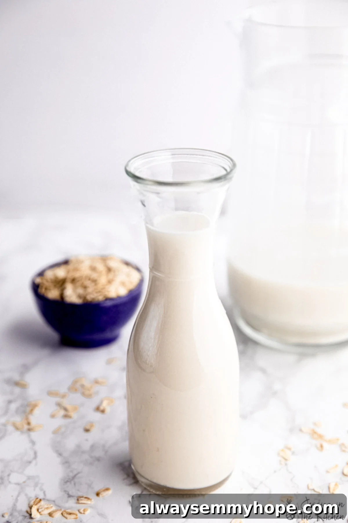 Discover this easy oat milk recipe to make rich, creamy, and delicious homemade oat milk for a fraction of the cost of store-bought. Perfect for all your plant-based needs! Freshly made homemade oat milk in a glass jar, with a bowl of rolled oats and a larger jug of oat milk in the background, showcasing its rich and creamy texture.
