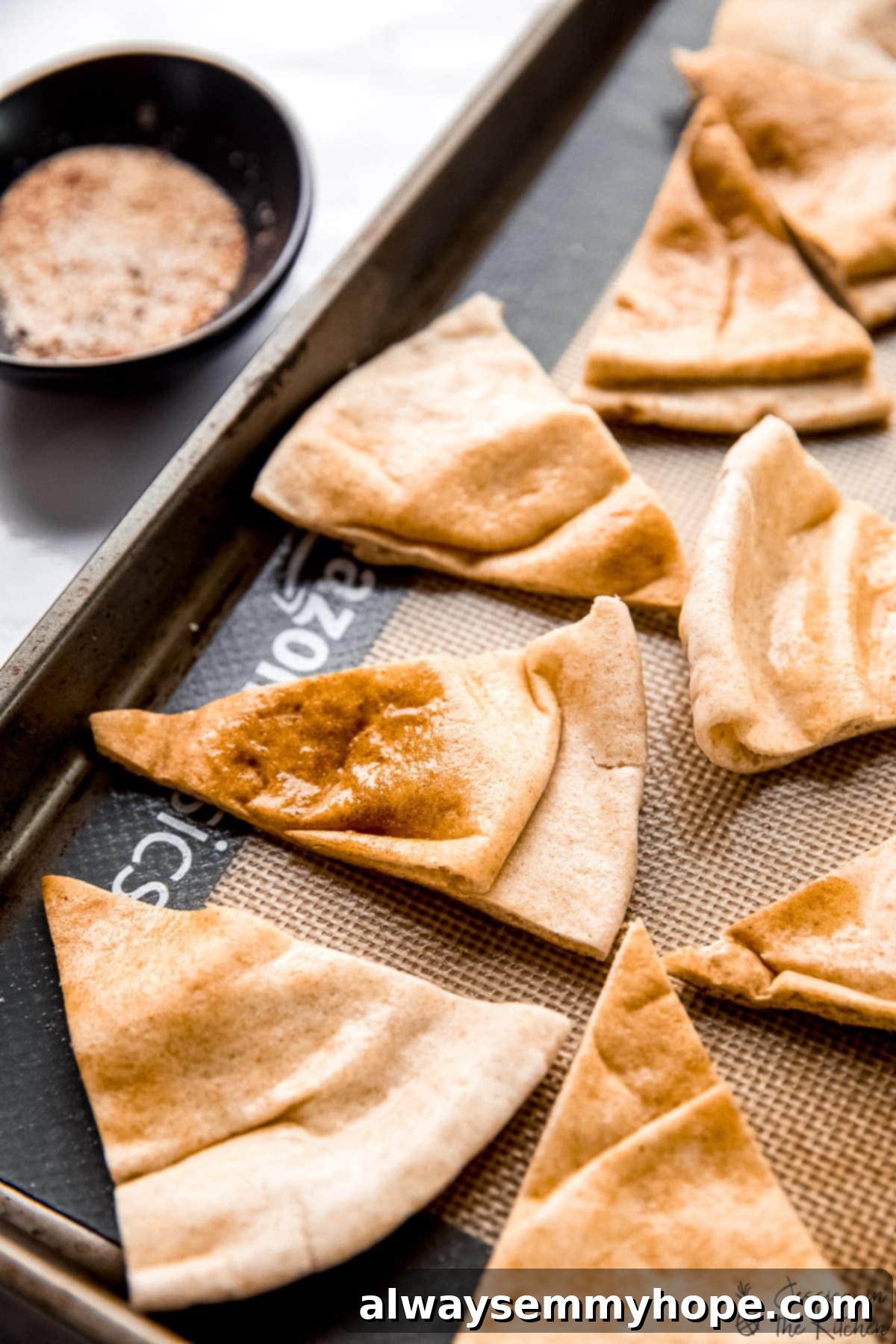 Pita bread triangles brushed with oil and seasoned, laid out on a baking sheet, with a bowl of seasoning mix beside it.