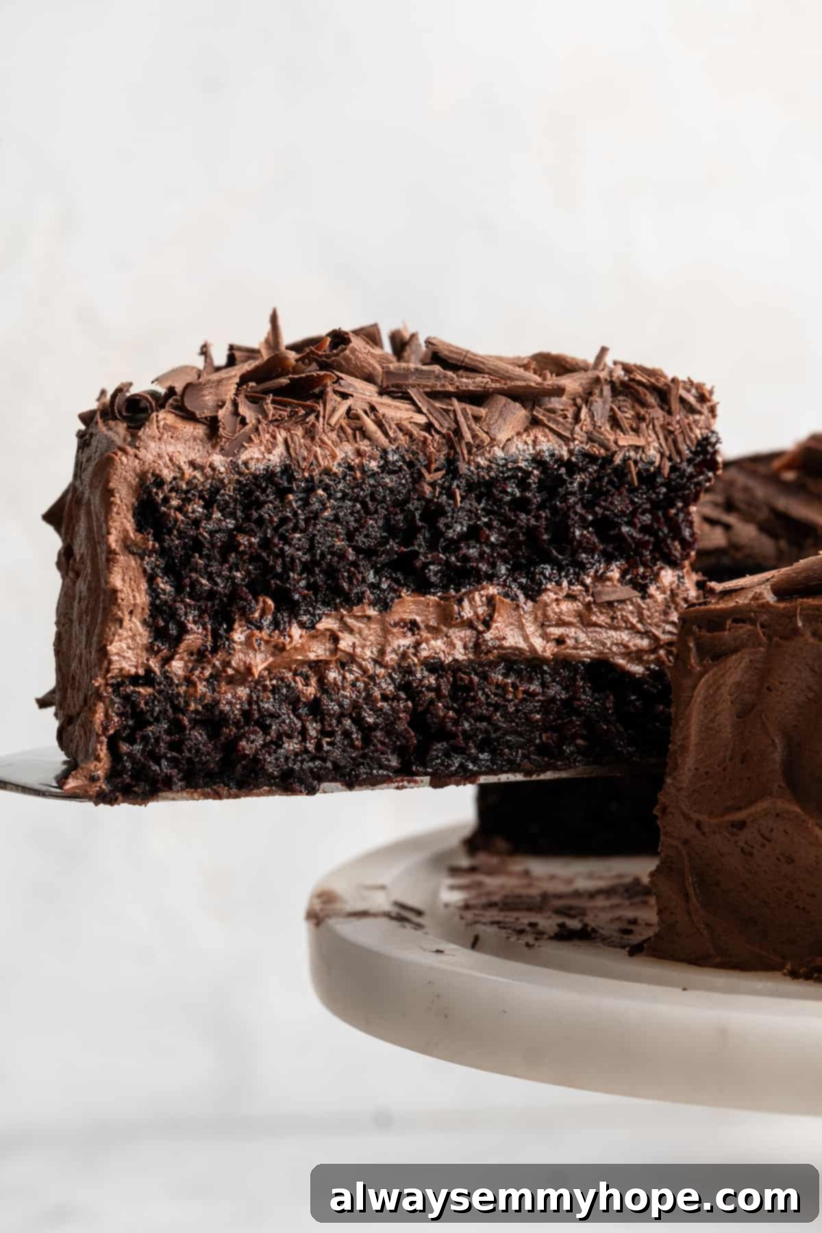 A slice of rich, moist vegan chocolate cake being removed from a cake stand, showcasing its perfect texture and chocolate frosting.