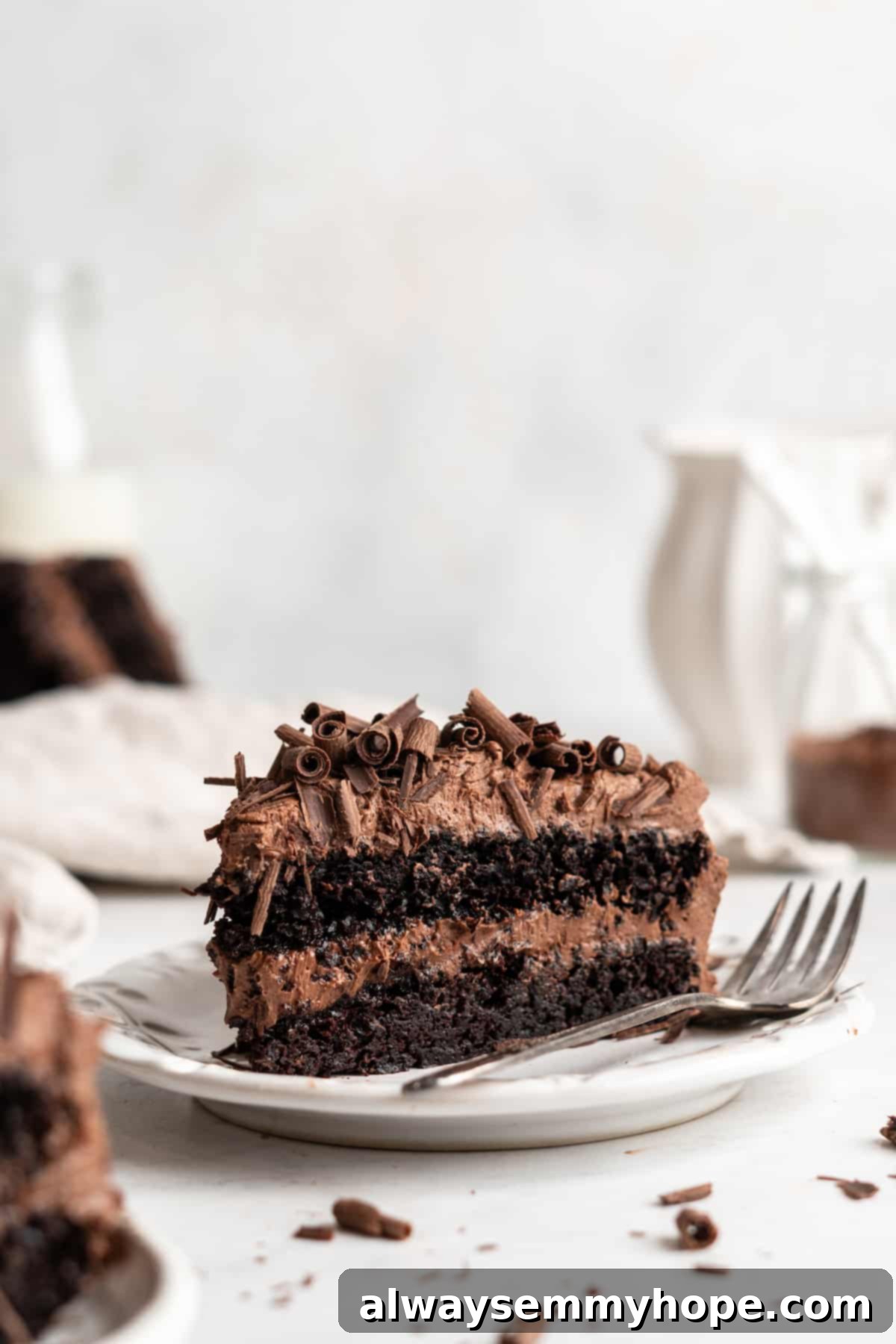 A close-up of a single slice of vegan chocolate cake on a plate with a fork, showing the fluffy cake texture and smooth frosting, ready to be enjoyed.