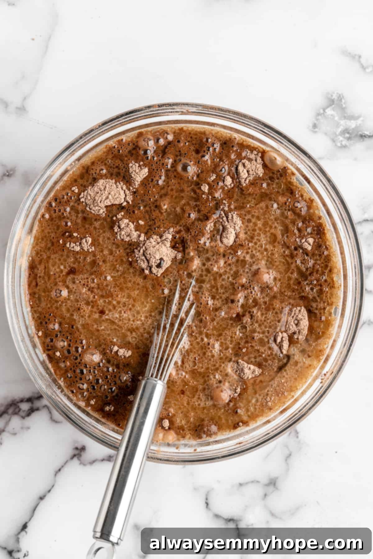 A whisk actively blending vegan chocolate cake batter in a clear glass bowl, showing a smooth, consistent mixture.