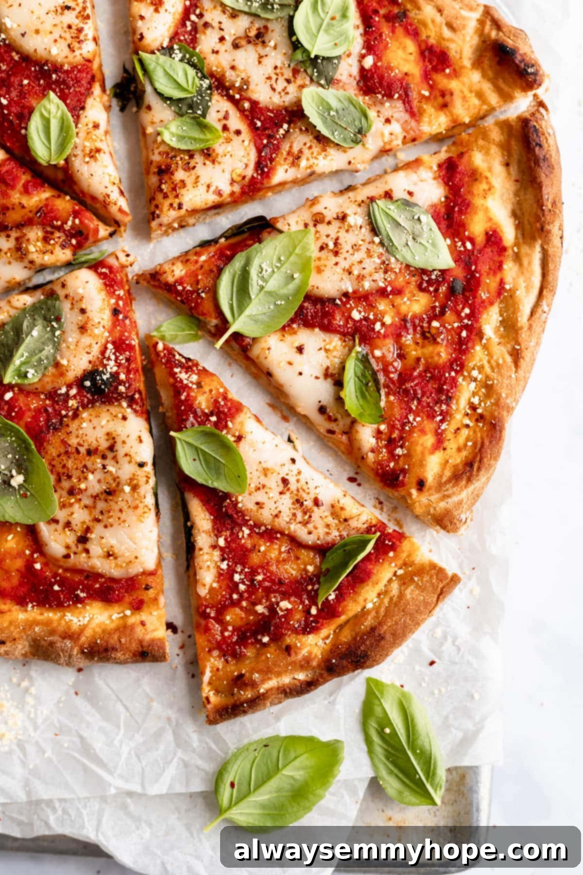 Overhead close-up of a freshly baked vegan pizza, sliced into wedges, with one slice gently pulled out. Fresh basil leaves are scattered around on parchment paper on a baking sheet.