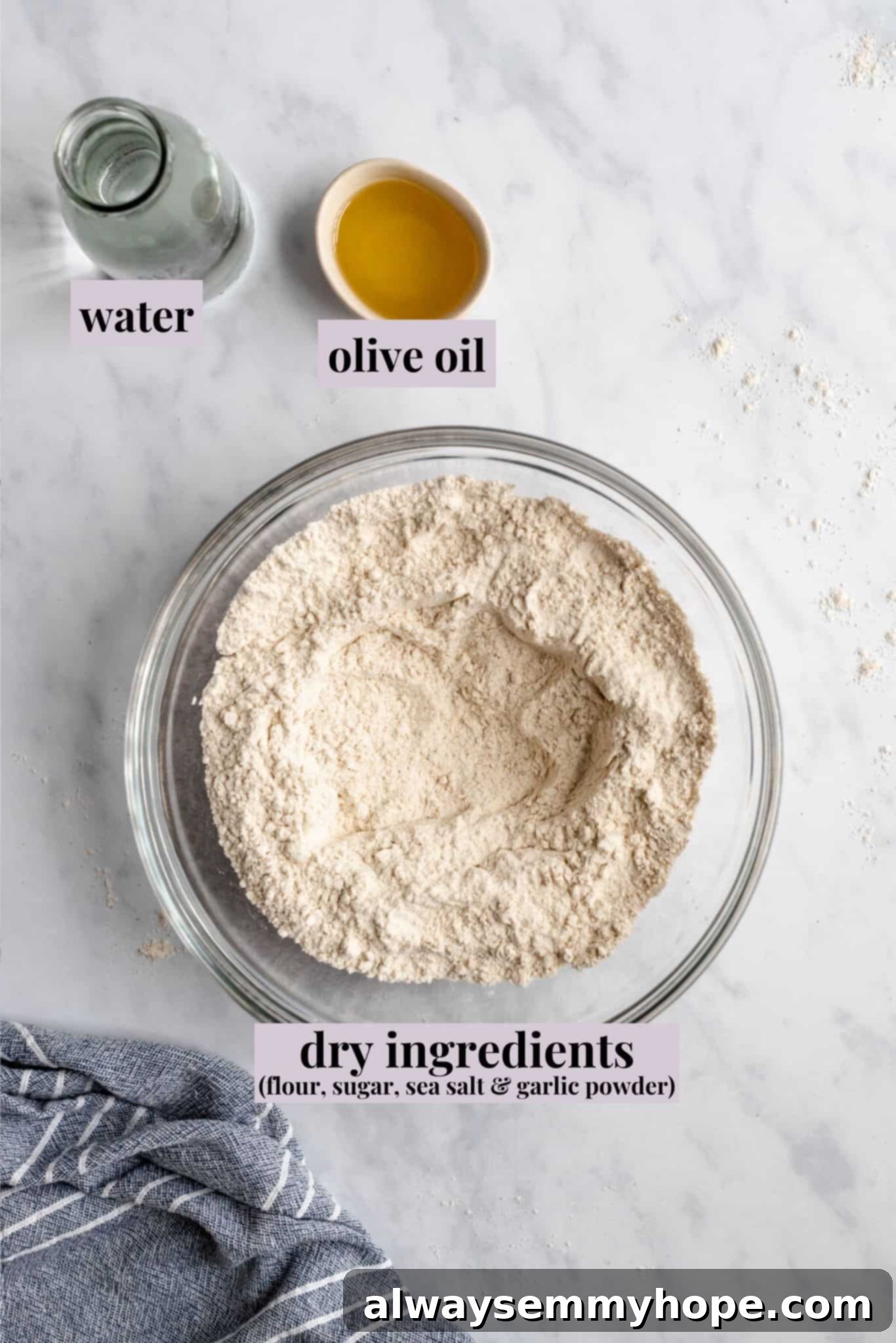 Close-up of various ingredients for vegan pizza dough neatly arranged on a white marble counter, including flour, olive oil, yeast, salt, sugar, and garlic powder.