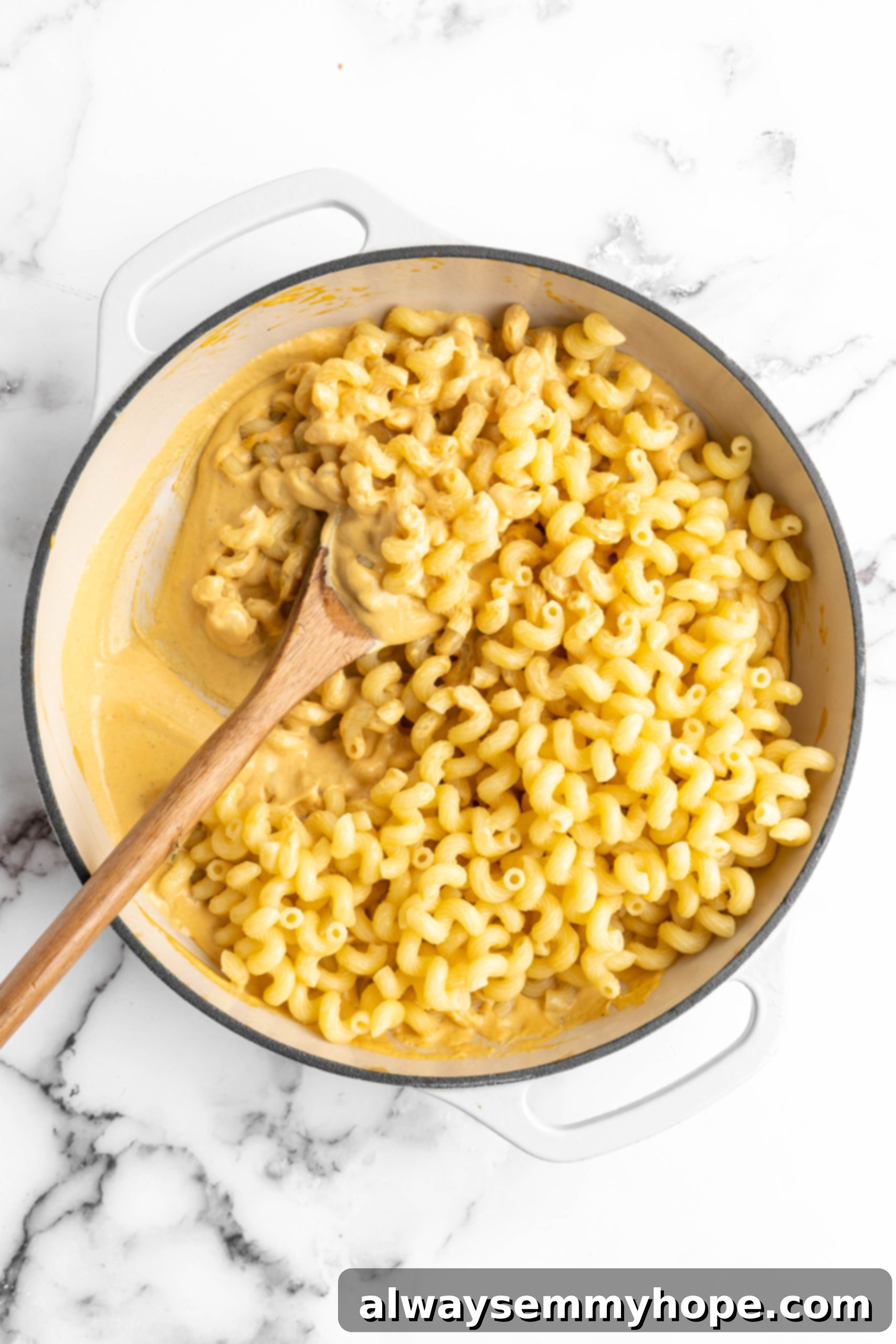 Cooked elbow macaroni pasta coated in rich vegan cheese sauce, stirred in a cast iron pan with a wooden spoon, ready for baking.