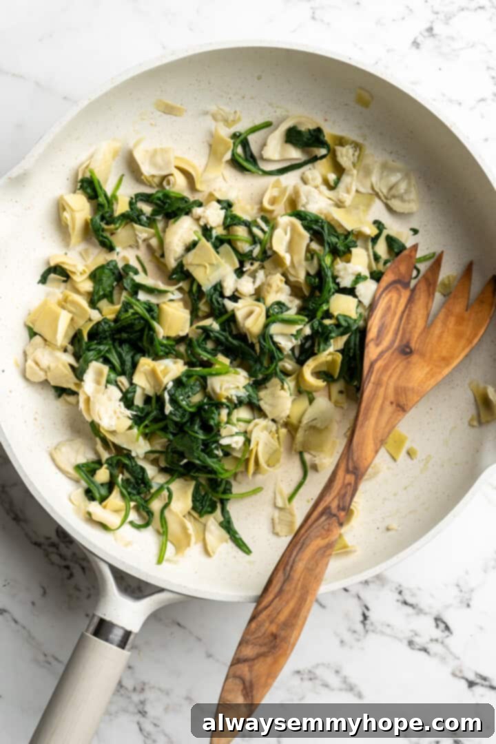 Spinach and artichokes combined in a pan, building the dip's foundation. Overhead view of wilted spinach and chopped artichoke hearts mingling in a skillet with a wooden spoon, creating a flavorful base.