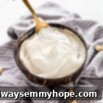 A spoon gracefully dips into a pristine white bowl filled with smooth, creamy homemade cashew cream, highlighting its luxurious texture.