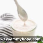 A delicate spoon drizzles luxurious vegan Alfredo sauce into a clear jar, highlighting its smooth and rich consistency.