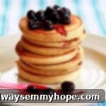 A beautifully stacked tower of golden vanilla pancakes, drizzled with sweet blueberry sauce, on a pristine white plate.