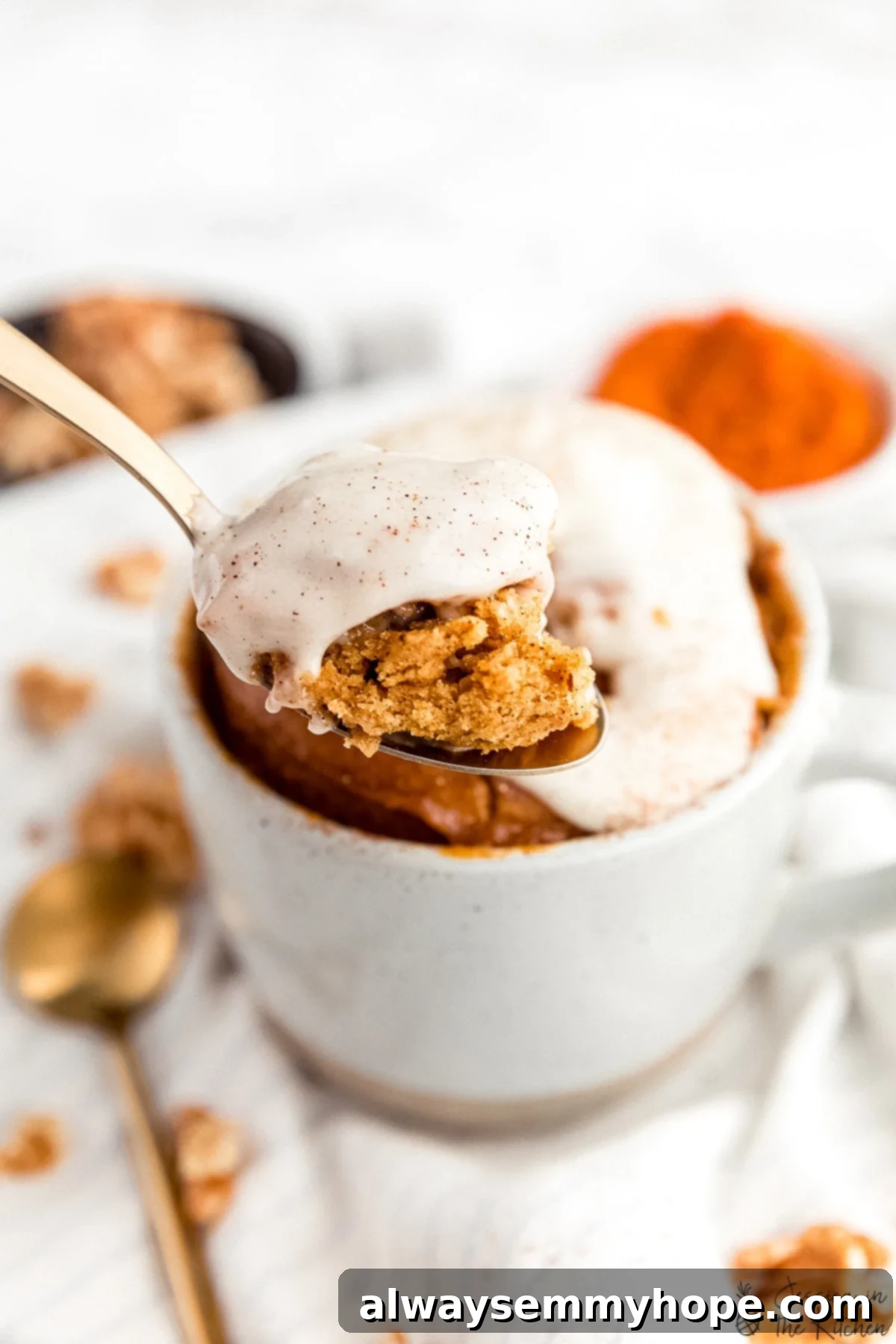 Spoon with frosted cake in front of a mug with pumpkin cake in it, highlighting the texture.