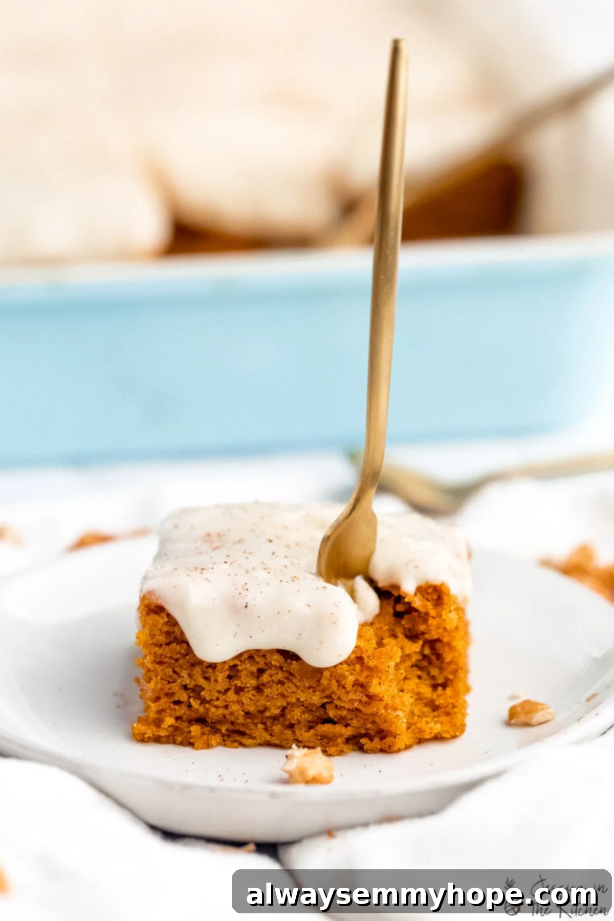 This Vegan Pumpkin Cake recipe is the perfect mix of moist, fluffy, and spiced. Top with cream cheese frosting for the ultimate fall dessert. Photo of a fork being stuck into a pumpkin cake on a plate with a bite missing in front of a blue container filled with slices of cake.