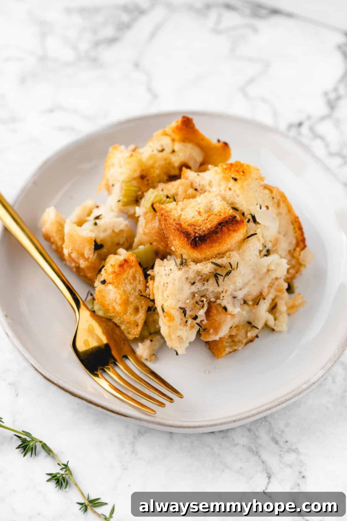 A perfectly scooped slice of vegan stuffing resting on a white plate, accompanied by a gold fork and fresh thyme sprigs, ready to be enjoyed.