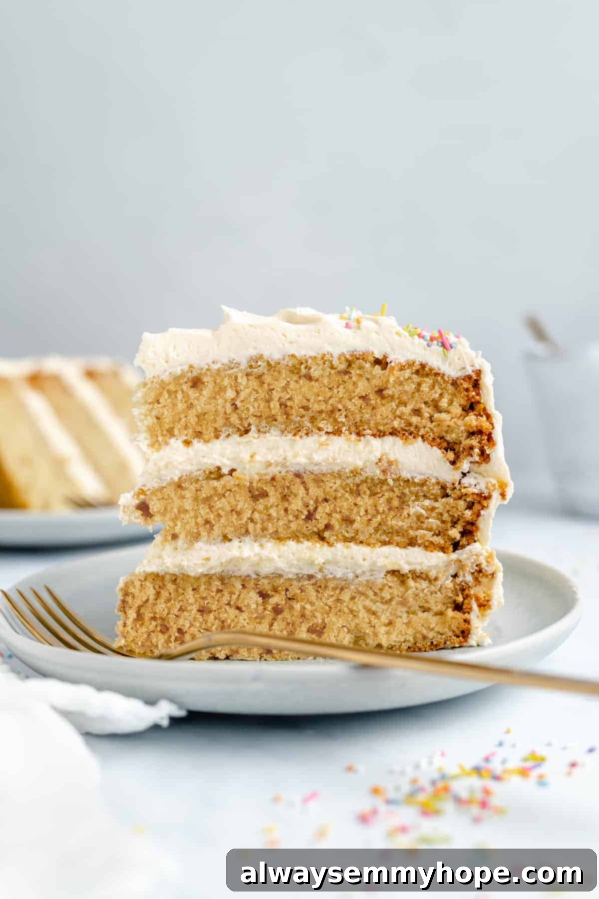 Classic Vegan Vanilla Cake with Buttercream Frosting 2 An upright slice of vegan vanilla cake on a white plate, showcasing its beautiful layers and elegant buttercream finish, with a golden fork in front.