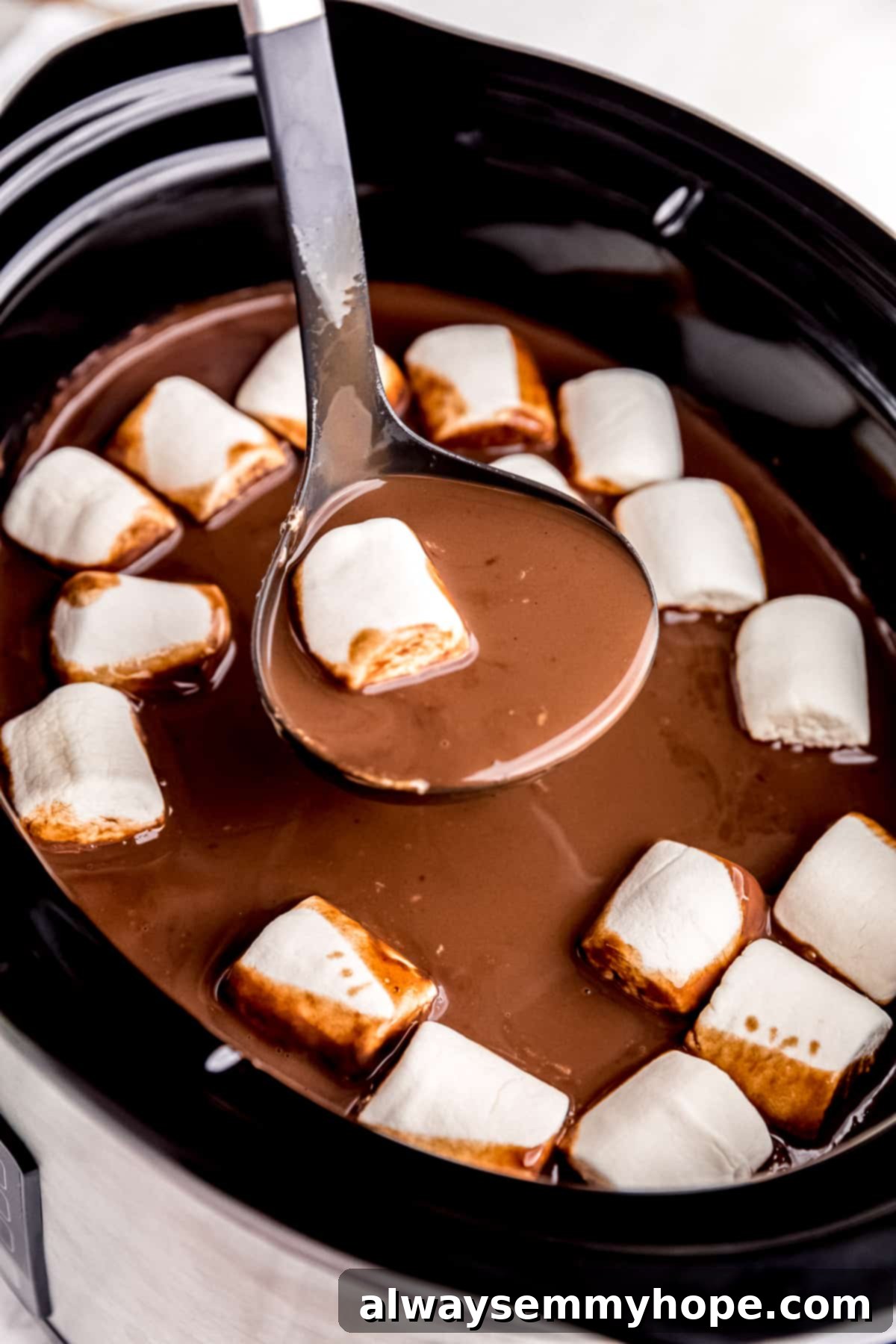 A delicious shot of slow cooker hot cocoa being served, complete with vegan marshmallows. A ladle serving slow cooker hot chocolate into a mug, garnished with vegan marshmallows and a sprinkle of cocoa