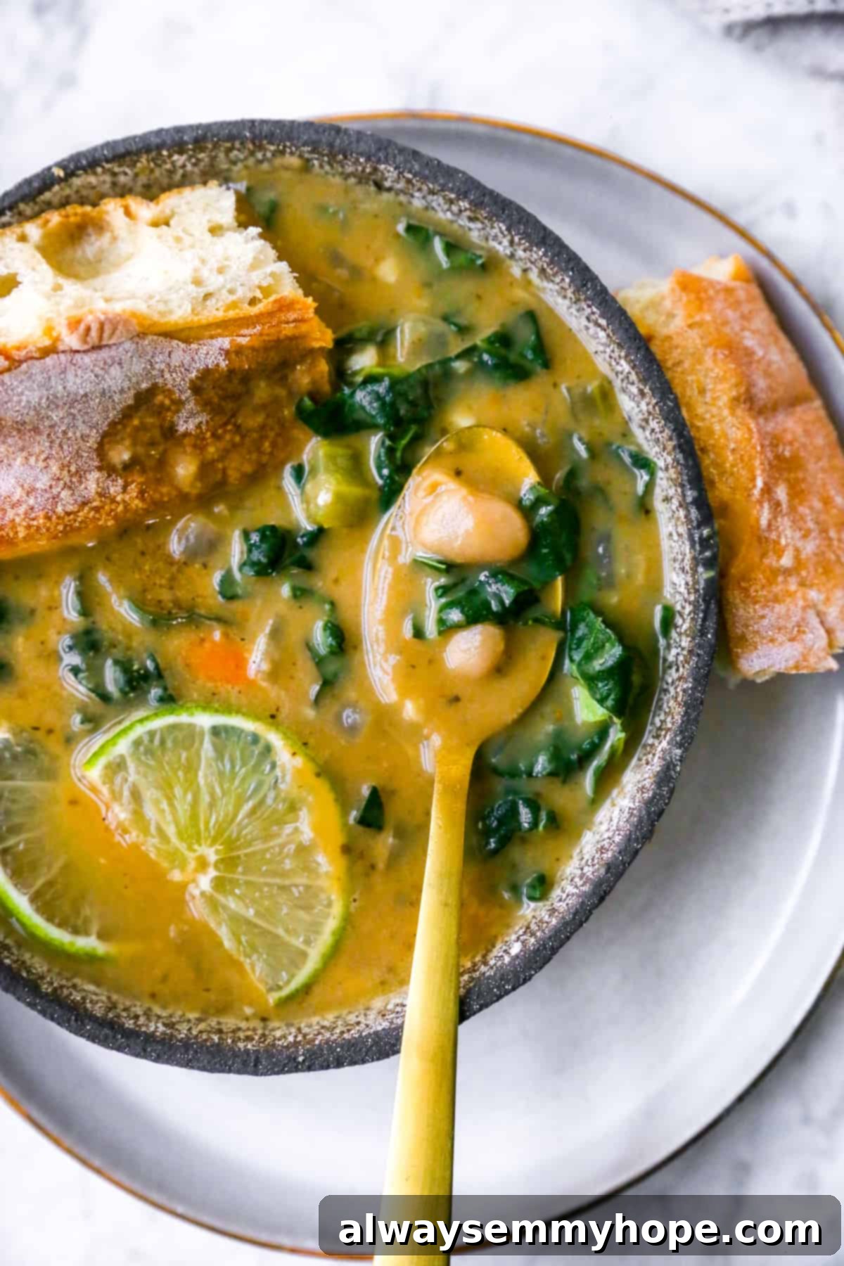 A comforting bowl of Tuscan White Bean Soup (Ribollita) with a gold spoon, accompanied by crusty bread, fresh lime, and vibrant kale. Perfect for a cozy meal.