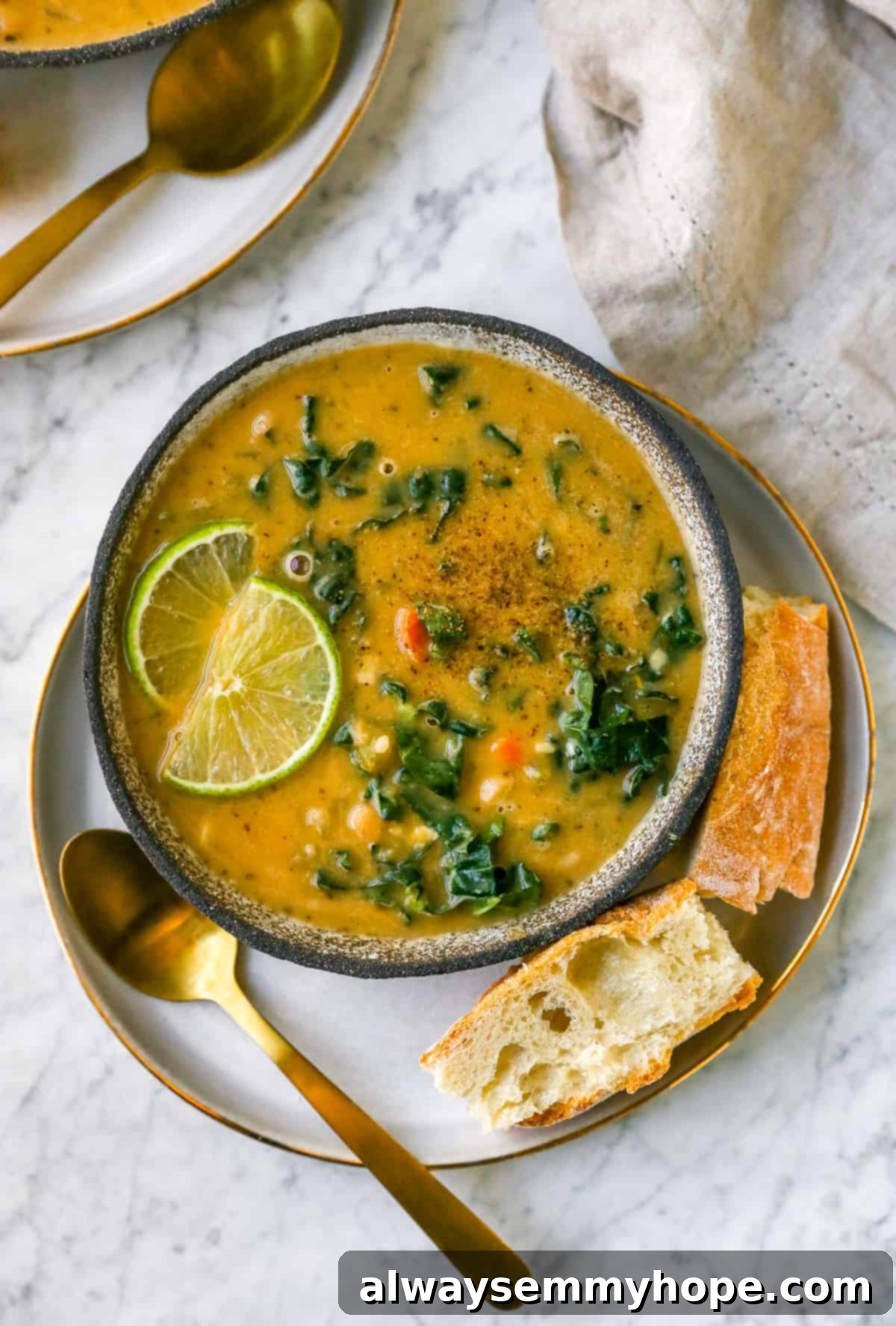 A beautifully presented bowl of Tuscan White Bean Soup on a plate, with a golden spoon, fresh bread, lime wedges, and kale garnish, ready to be served.