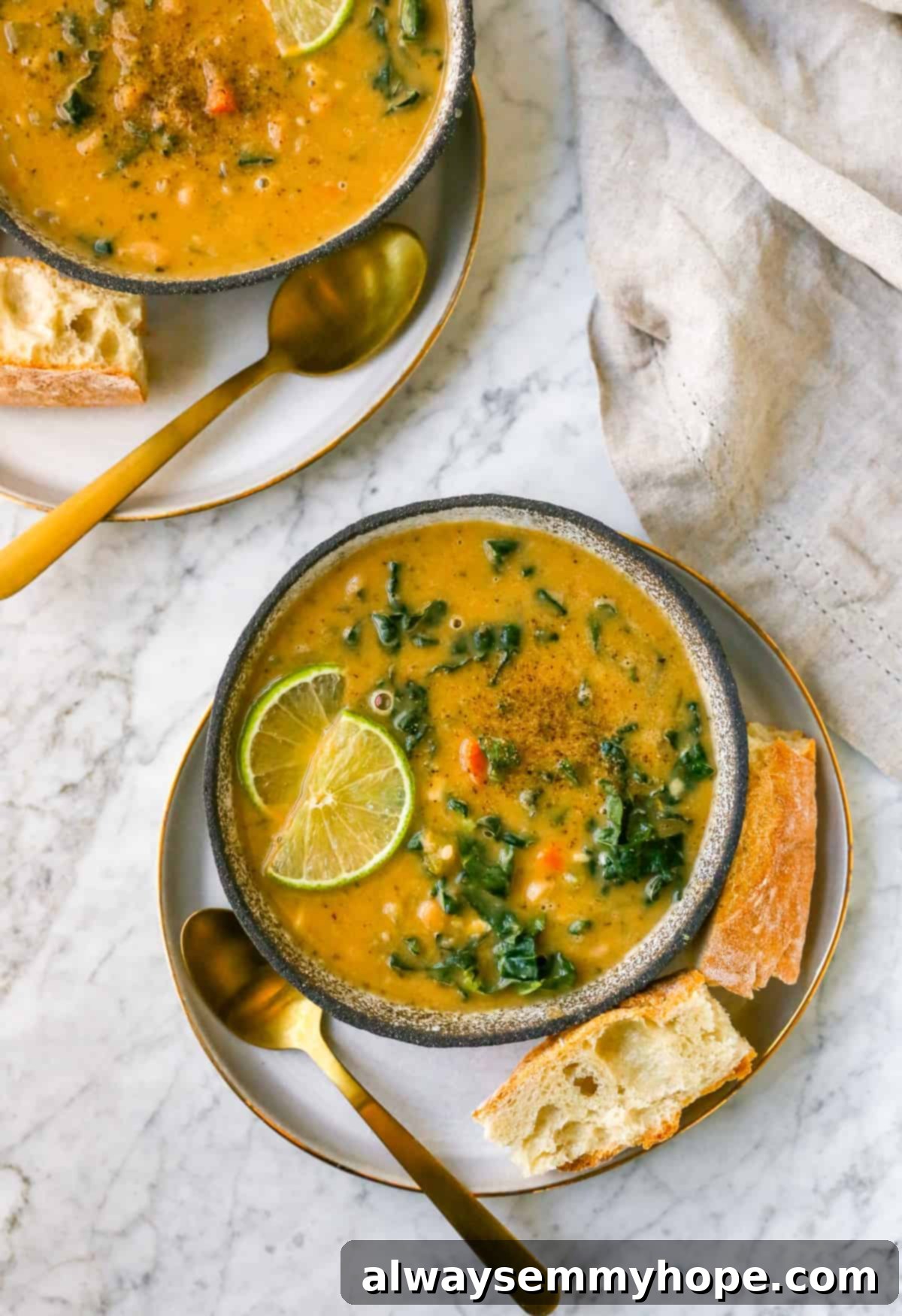 A closer look at a serving of Tuscan White Bean Soup (Ribollita) on a plate, garnished with a gold spoon, fresh bread, lime wedges, and green kale leaves.