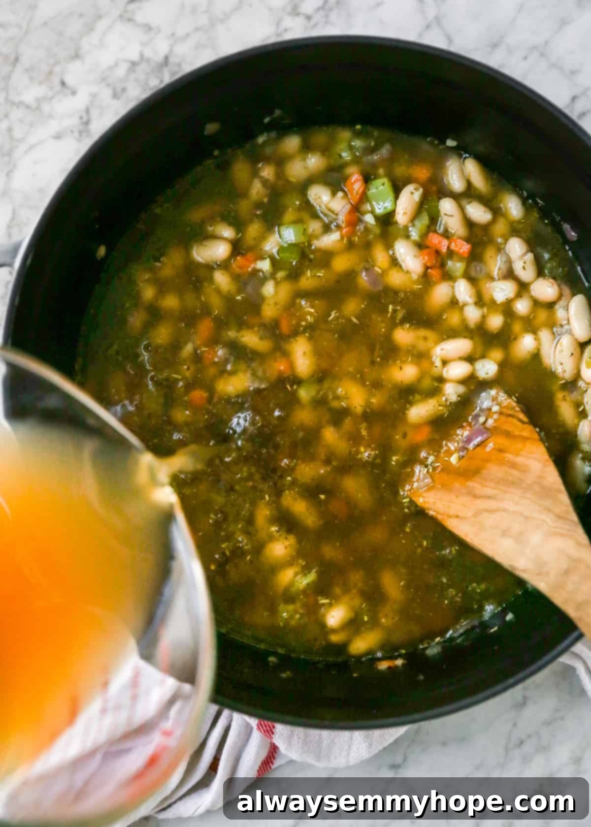 Pouring vegetable broth into the pot with sautéed vegetables, beans, and seasoning, ready for simmering to create a flavorful white bean soup.