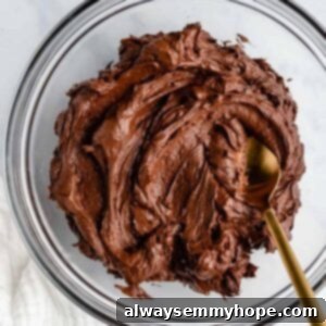 Glass mixing bowl of rich chocolate frosting with a gold spoon