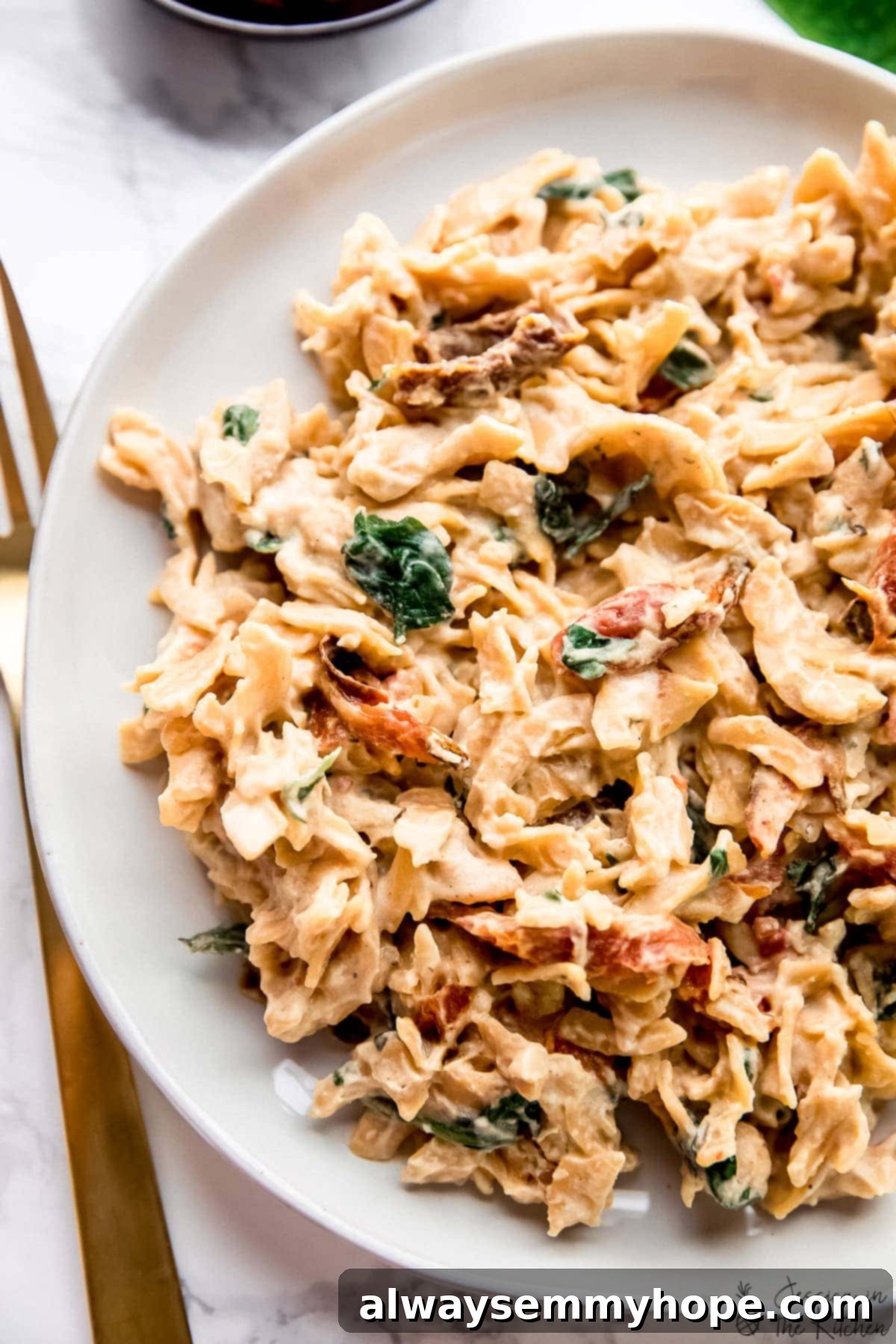 Pasta tossed with hummus, spinach, and sun dried tomatoes on a white plate, ready to be enjoyed as a quick vegan meal.