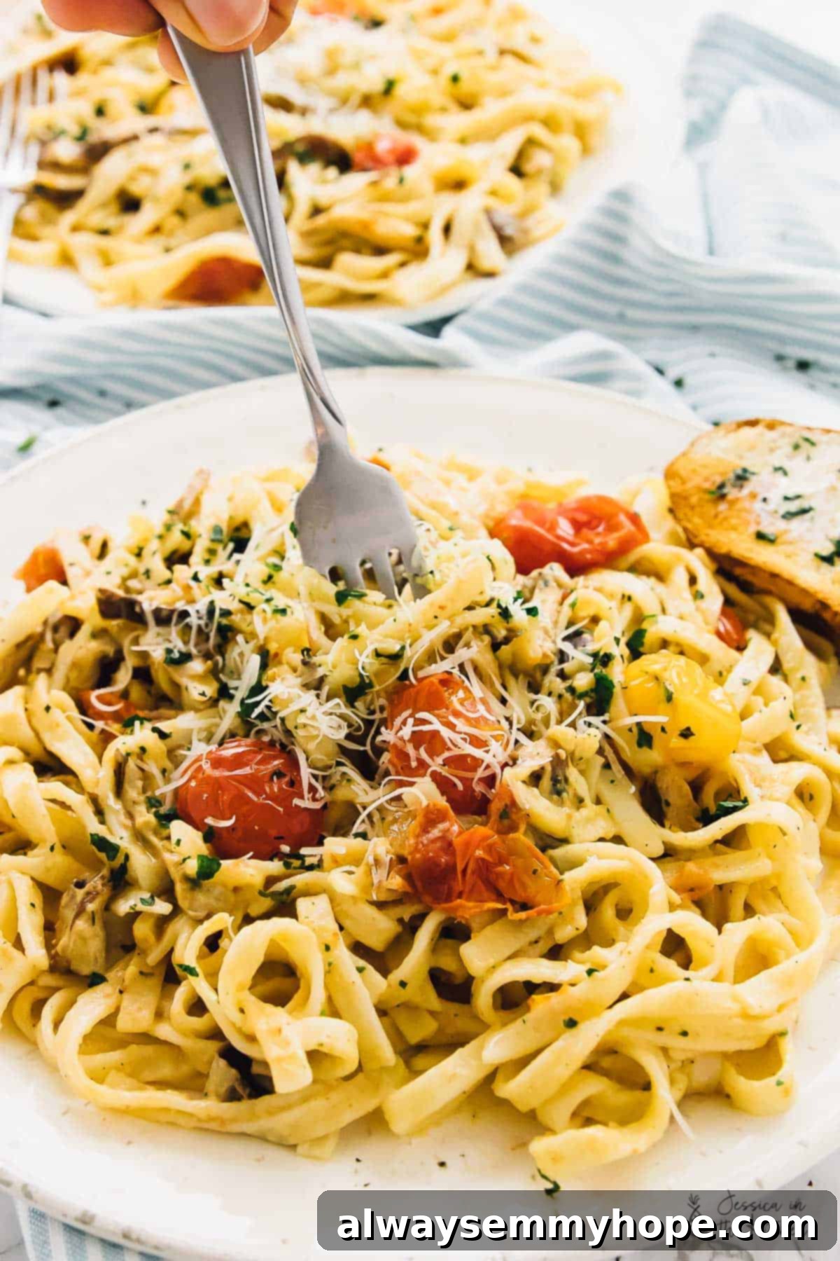 A fork lifts a mouthful of fettuccine pasta, adorned with burst tomatoes, fresh herbs, and a sprinkle of vegan Parmesan cheese.