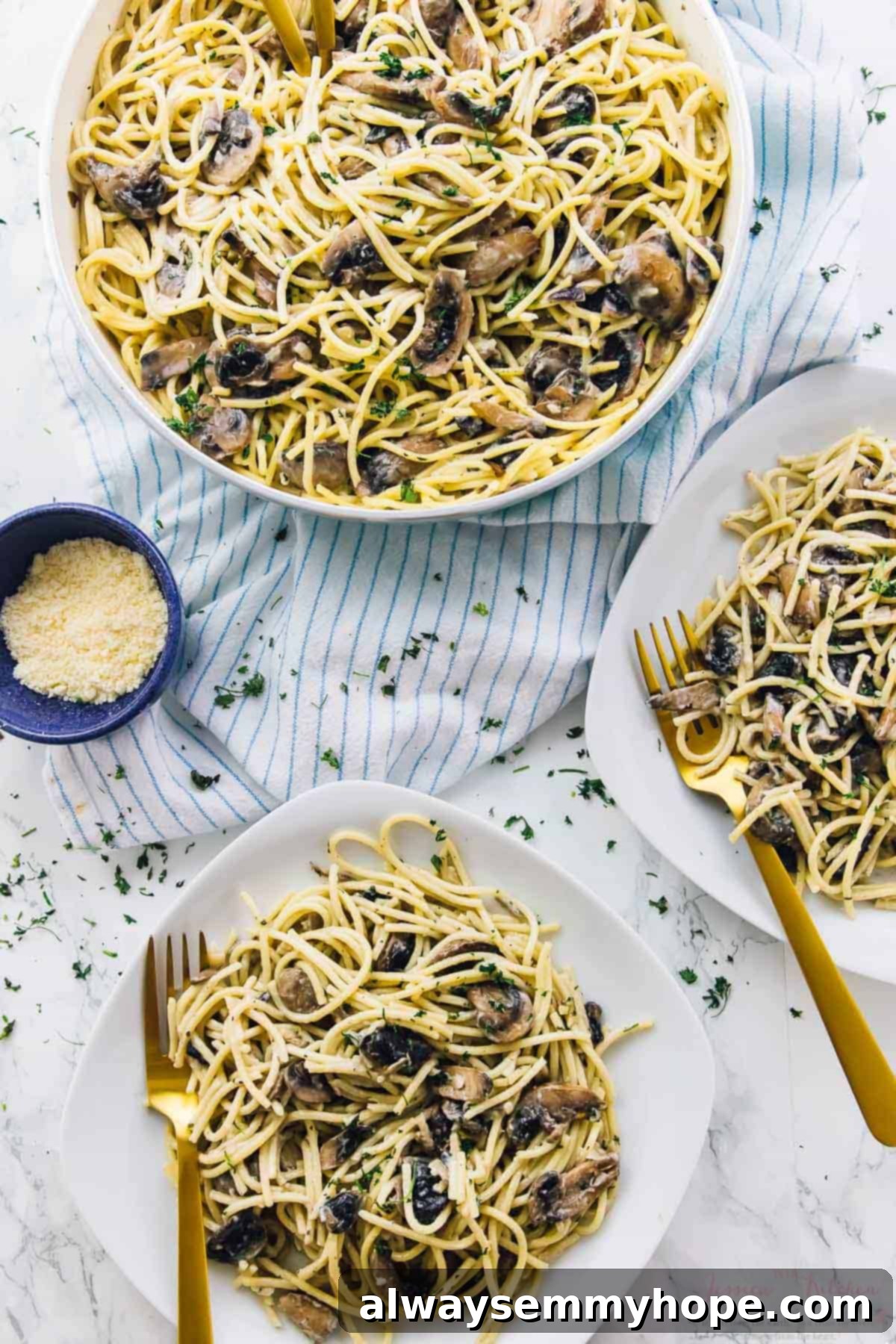 Two plates of creamy mushroom vegan alfredo pasta, garnished with fresh herbs, alongside gold forks and a bowl of vegan parmesan cheese.