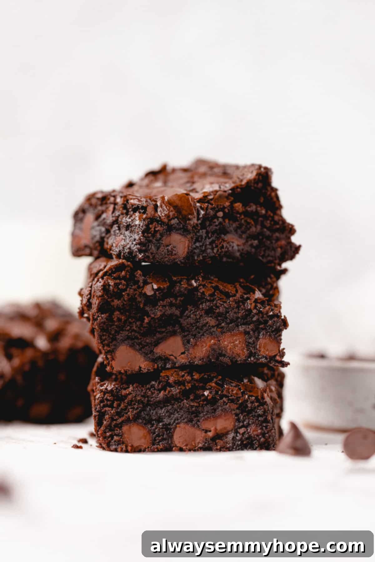 three fudgy gluten-free vegan brownies stacked on top of one another with one in the corner and a bowl of chocolate chips