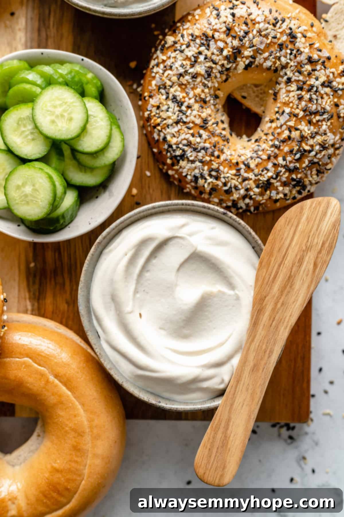 Creamy plain vegan cream cheese spread on a bagel, with other bagels and ingredients in the background on a wooden cutting board.