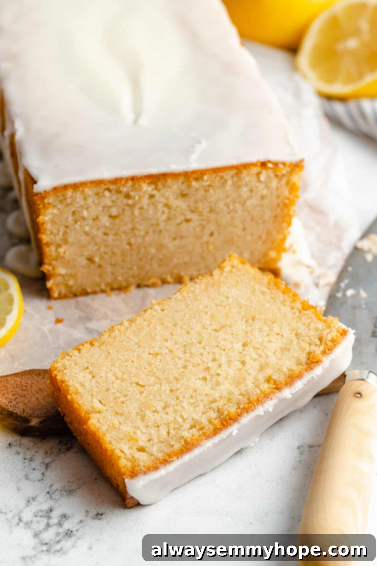 A slice of vegan lemon pound cake with a generous drizzle of lemon glaze, resting on a rustic wooden surface next to the full loaf.