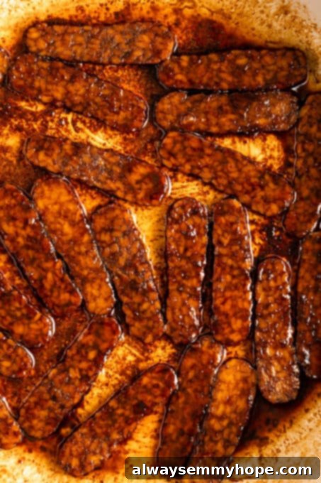A close-up shot of thinly sliced tempeh bacon sizzling in a hot pan, showing the crispy edges and savory marinade. Tempeh is a fermented soy product that makes an excellent, protein-packed vegan meat alternative.