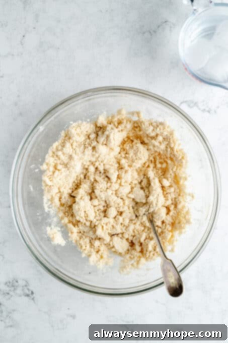 The coarse pie dough mixture in a mixing bowl, with ice cold water being slowly added to bring it together.