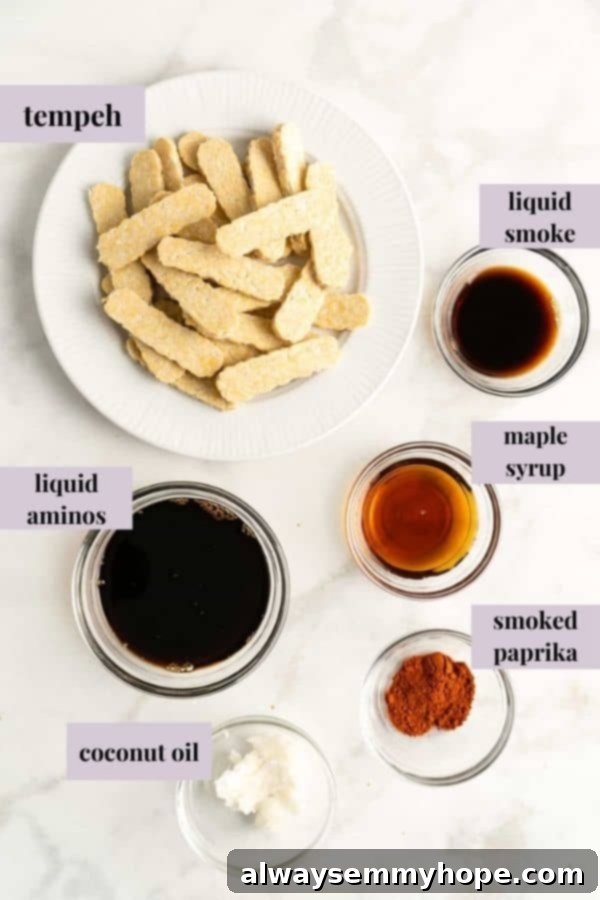Overhead view of all the ingredients for tempeh bacon laid out on a dark surface, including tempeh, soy sauce, liquid smoke, and maple syrup.