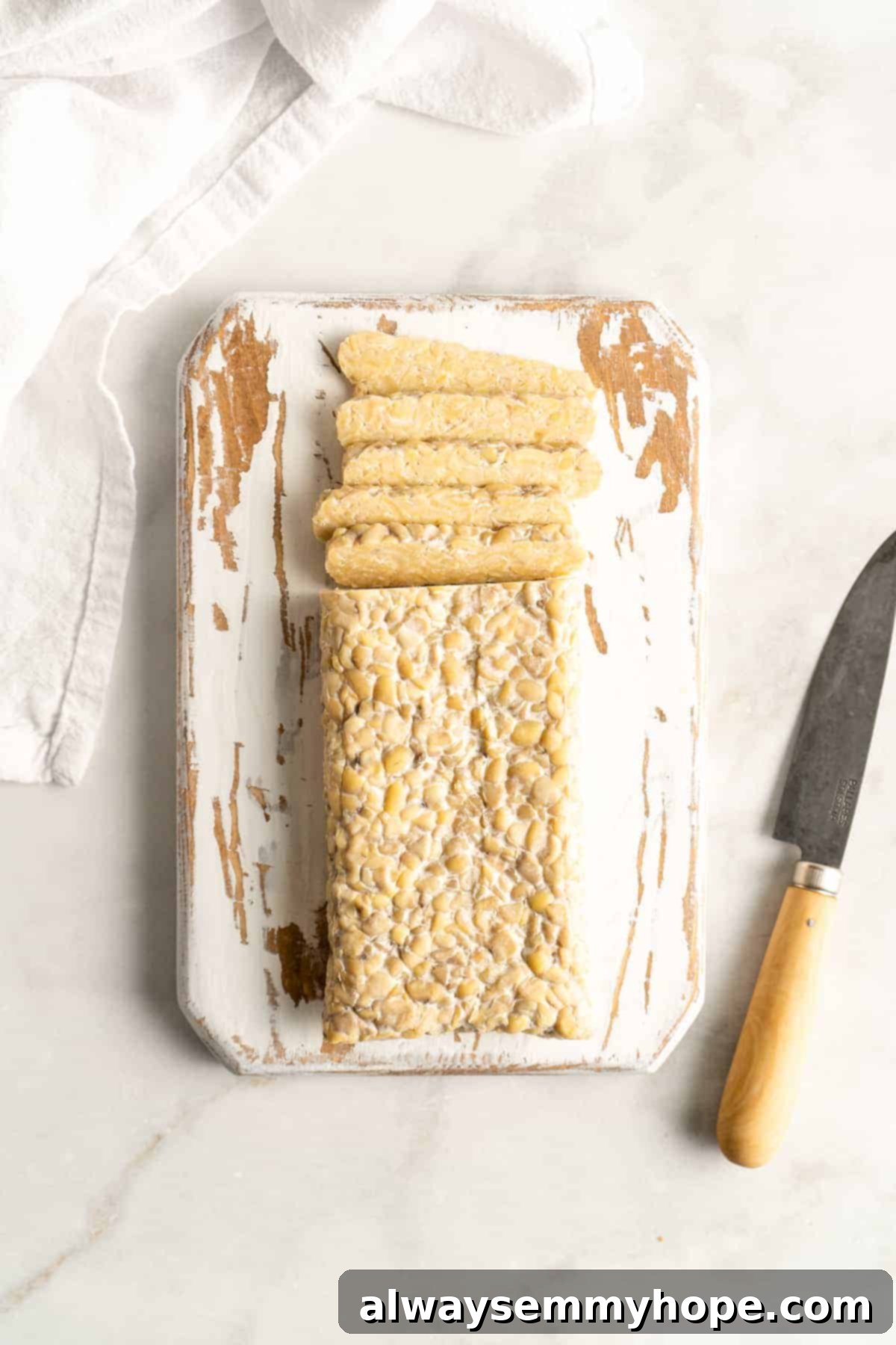 Overhead shot of tempeh being sliced on a rustic white painted cutting board, preparing for cooking.