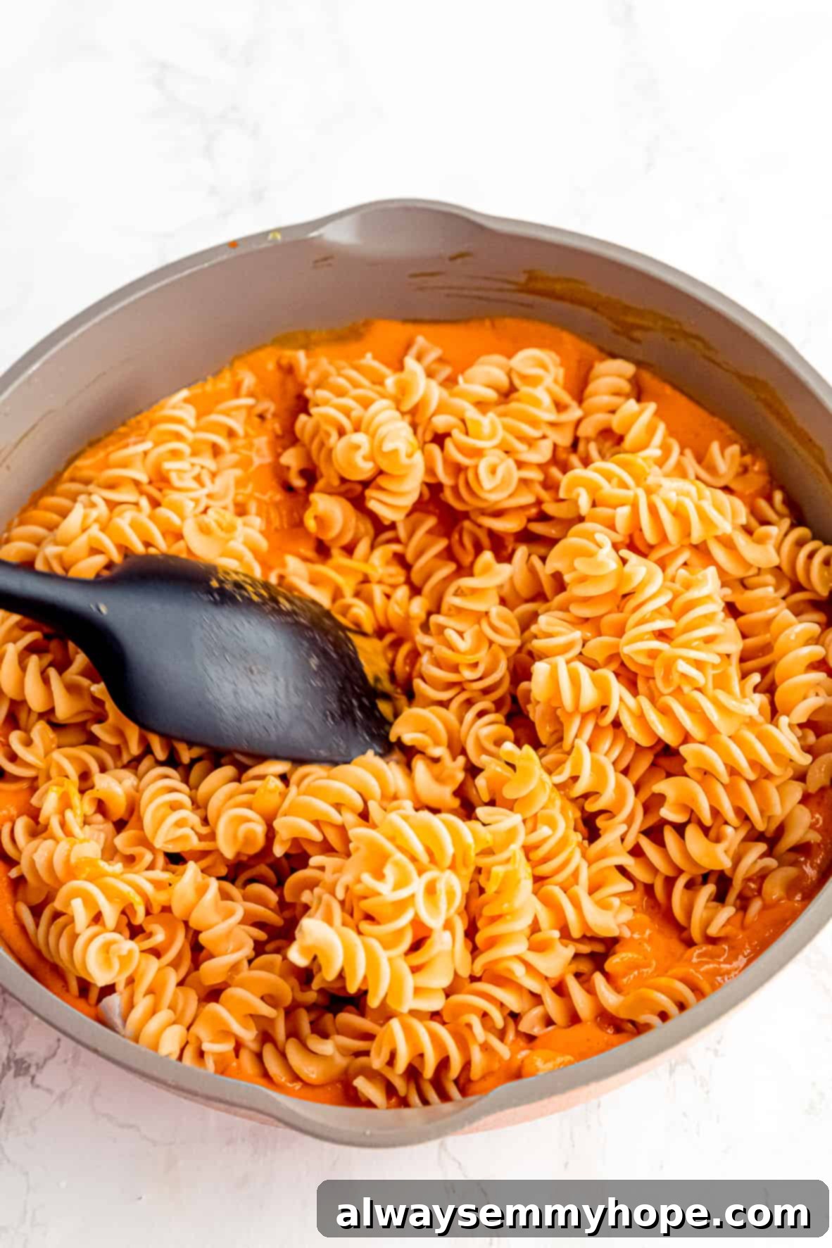 A spatula stirring cooked pasta into roasted red pepper pasta sauce in a skillet.