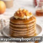 A towering stack of fluffy apple pie pancakes on a plate, adorned with a dollop of yogurt, maple syrup, and tender spiced apple slices on top.
