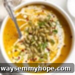 Overhead view of creamy vegan pumpkin soup in a bowl with a spoon, garnished with crunchy pepitas, fresh green onions, and a delicate swirl of coconut cream.