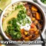 Overhead view of a hearty sweet potato and black bean chili served in a bowl, generously garnished with creamy cashew sour cream, diced avocado, fresh cilantro, and vibrant green onion.