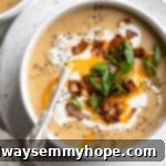 Overhead view of a comforting bowl of vegan loaded potato soup, generously topped with shredded vegan cheese, a dollop of plant-based yogurt, crispy vegan bacon bits, and freshly chopped scallions.