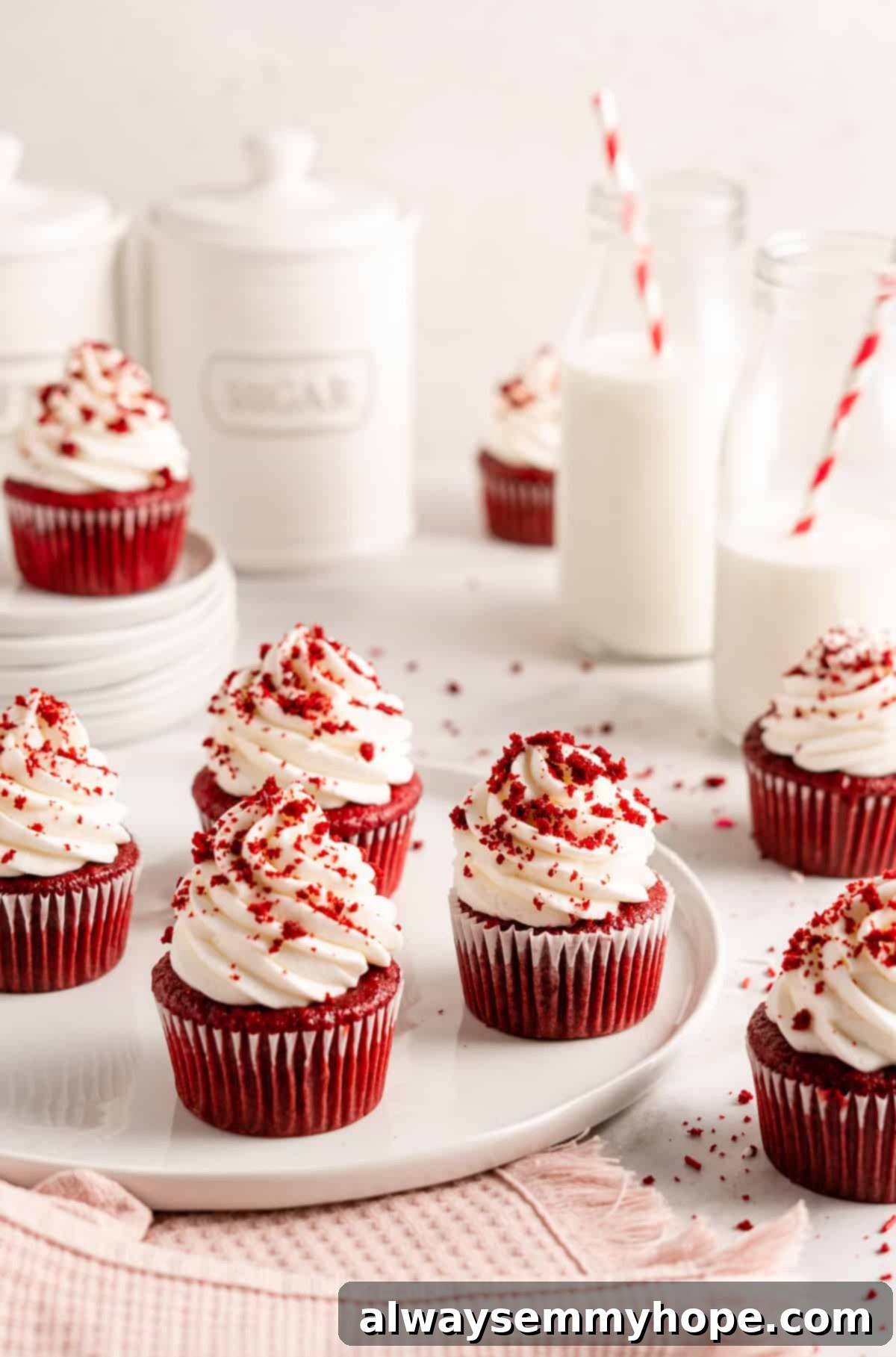 A trio of freshly baked vegan red velvet cupcakes, richly decorated with piped swirls of vegan cream cheese frosting and a scattering of vibrant cake crumbs, presented on a light plate.