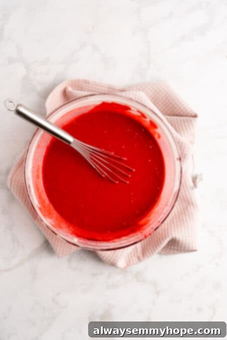 Overhead view of a glass mixing bowl containing vibrant red velvet cake batter being whisked by an electric mixer, showing its smooth and well-combined consistency.