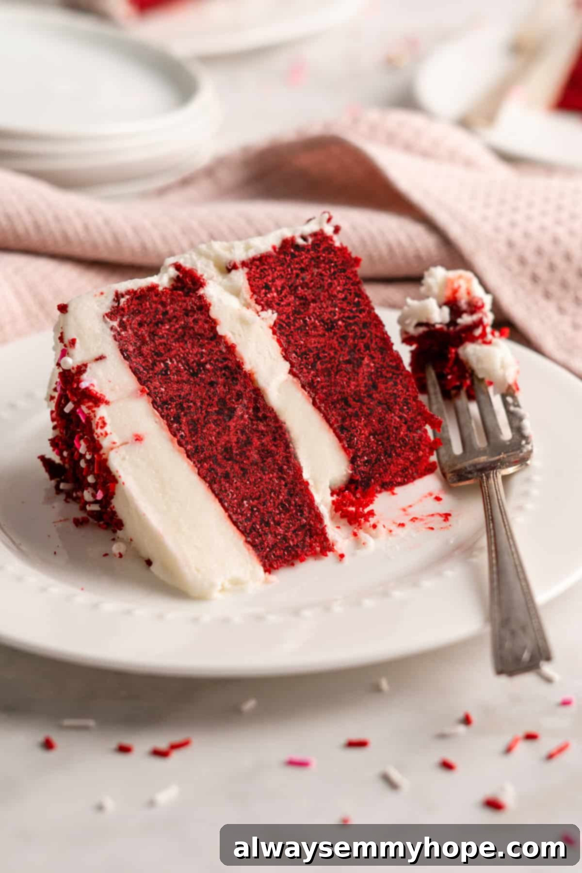A decadent slice of vegan red velvet cake on a white plate with a fork, showing off its rich red color and thick layer of cream cheese frosting.